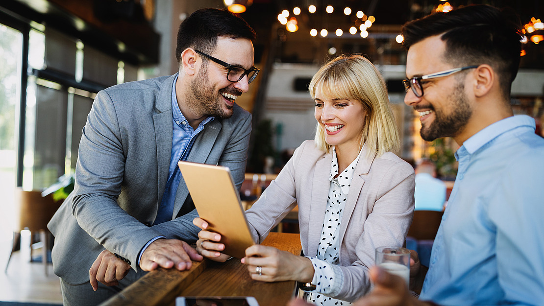 Drei Kollegen diskutieren fröhlich etwas auf einem Tablet in einem modernen Büroraum. Eine Frau mit blondem Haar hält das Tablet, während zwei Männer mit Brillen, alle in Business-Kleidung, mit ihr reden.