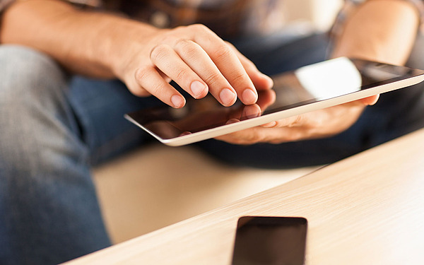 Nahaufnahme einer Person, die mit einer Hand ein Tablet verwendet und es mit der anderen hält. Die Person sitzt, trägt Jeans und ein Smartphone liegt auf einem Holztisch in der Nähe.