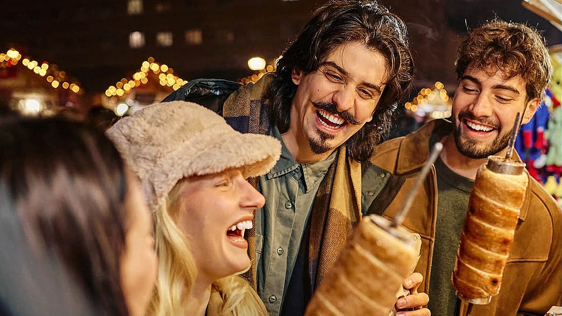 Vier Freunde, die auf einem Nachtmarkt lachen, während sie traditionelle Schornsteinbrötchen in den Händen halten.