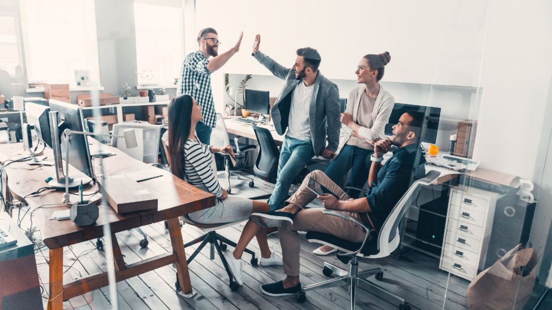 Eine Gruppe von fünf jungen Erwachsenen in einem modernen Büro, die sich locker unterhalten und High Fives geben. Einige sitzen auf Stühlen, andere stehen. Der Arbeitsbereich ist mit Schreibtischen, Computern und Büromaterial ausgestattet.