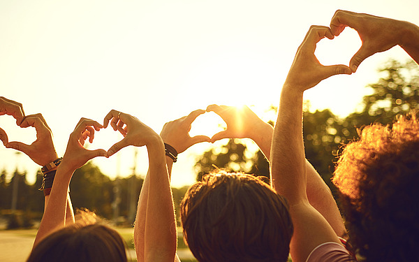Eine Gruppe von Menschen im Freien hält ihre Hände in Form von Herzen hoch, die sich als Silhouette vor der Sonne und einem unscharfen Hintergrund aus Bäumen abheben und so Einheit und Liebe zum Ausdruck bringen.