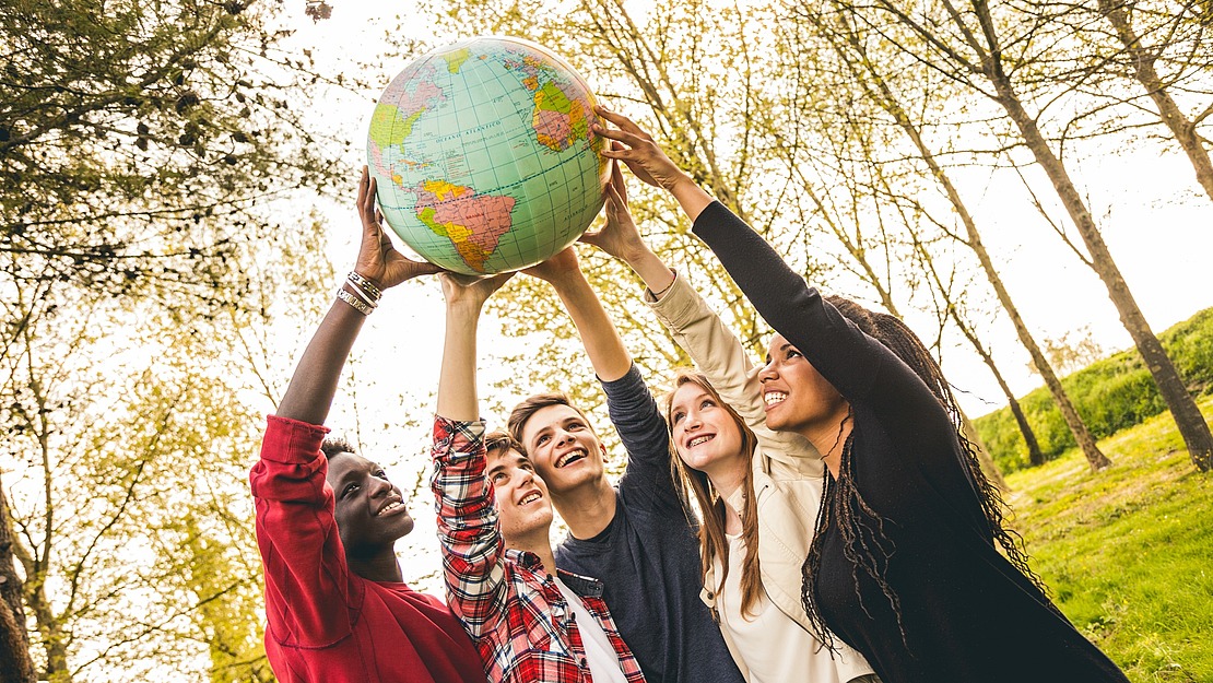 Vier lächelnde junge Erwachsene stehen im Freien und halten gemeinsam eine große Weltkugel, umgeben von Bäumen und Sonnenlicht.