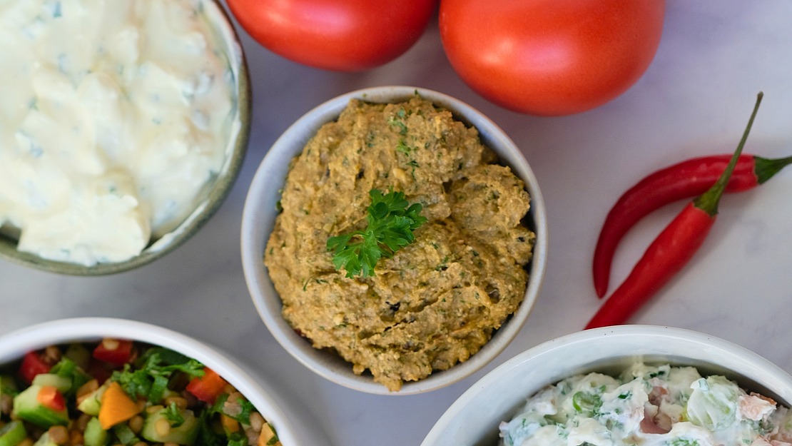 Schalen mit verschiedenen Dips und Salaten stehen auf einem Tisch, umgeben von frischen Tomaten und roten Chilischoten. Die Gerichte enthalten cremige und stückige Texturen, garniert mit Kräutern und Gemüse.