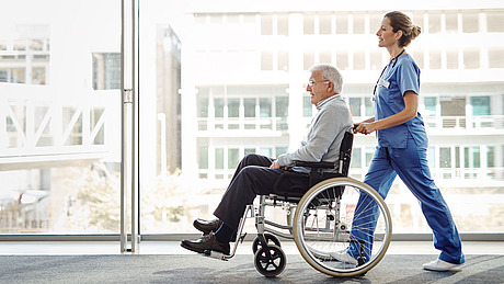 Eine Krankenschwester schiebt einen älteren Mann im Rollstuhl einen Flur entlang.