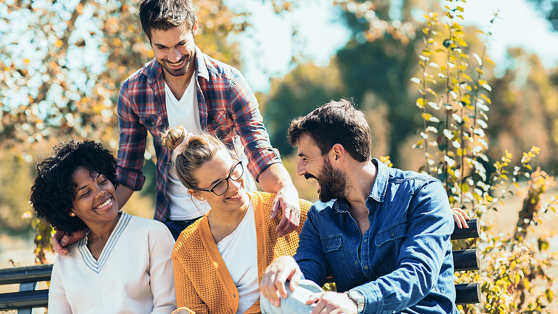 Vier Freunde sitzen an einem sonnigen Tag zusammen auf einer Gartenbank und lächeln und lachen. Zwei Frauen sitzen vor ihnen, eine mit Locken und eine mit Brille, während zwei Männer hinter ihnen stehen und sitzen. Alle wirken glücklich und entspannt.