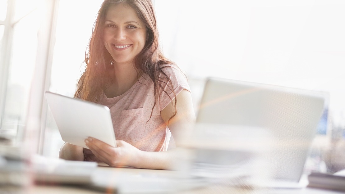 Eine lächelnde Frau sitzt an einem Schreibtisch und hält ein Tablet in der Hand, vor ihr liegen ein geöffneter Laptop und Papiere. Das Sonnenlicht fällt durch große Fenster im Hintergrund und schafft einen hellen, einladenden Arbeitsbereich.