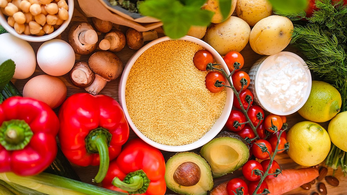 Eine bunte Mischung frischer Zutaten, darunter rote Paprika, Avocados, Kirschtomaten, Eier, Pilze, Zitronen, Kartoffeln, Kichererbsen, Hüttenkäse und eine Schüssel Couscous, die auf einer Holzplatte präsentiert werden.