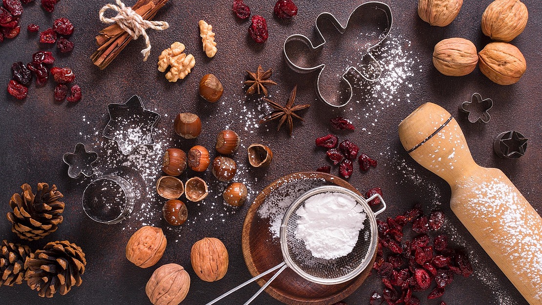 Flat-Lay-Aufnahme von Backzubehör, darunter Walnüsse, Haselnüsse, Cranberries, Zimtstangen, Ausstechformen, ein Nudelholz und ein mit Puderzucker gefülltes Sieb.