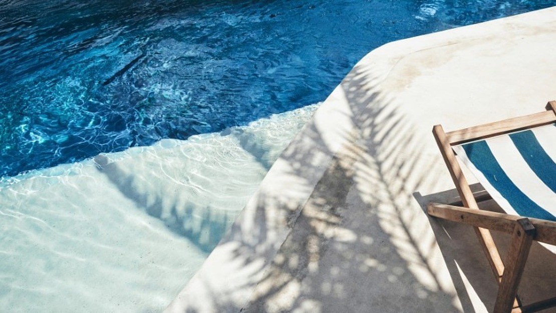 Ein blau-weiß gestreifter Liegestuhl steht auf einer sonnenbeschienenen Poolterrasse, auf der klares blaues Wasser und der Schatten einer Palme zu sehen sind.
