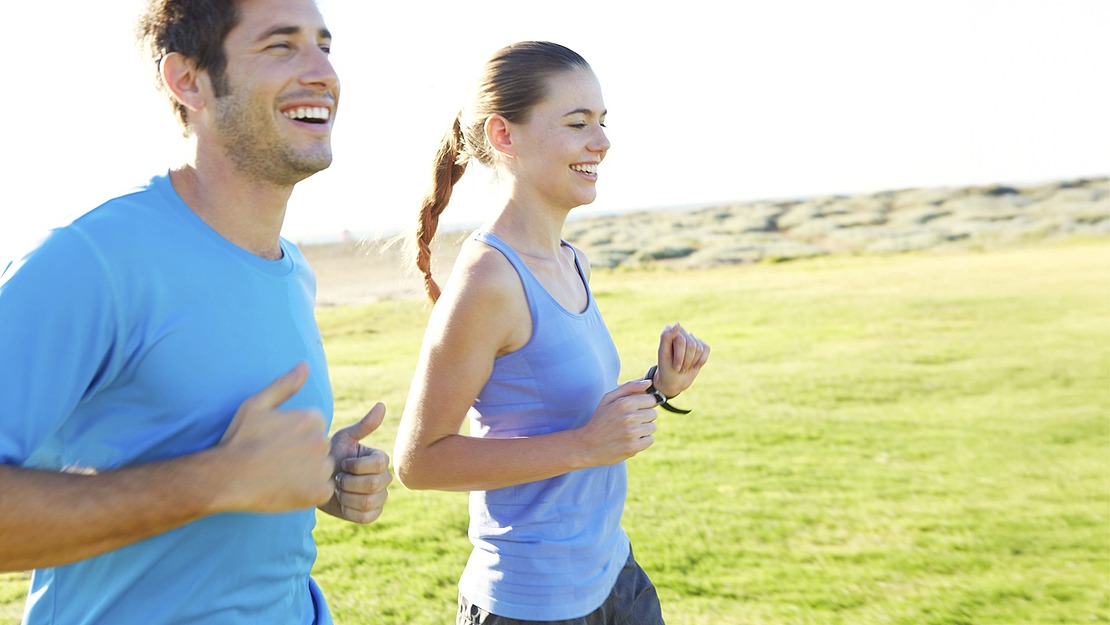 Ein Mann und eine Frau joggen Seite an Seite auf einer Wiese unter strahlendem Himmel, beide lächeln und tragen blaue Sportshirts.