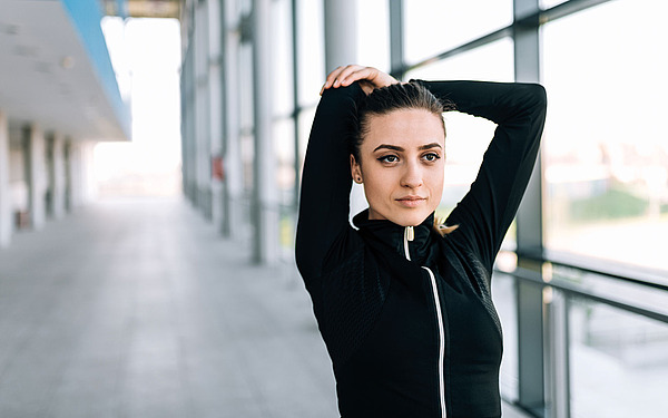 Eine Frau in Sportkleidung streckt ihren Arm über den Kopf, während sie in einem geräumigen Innenkorridor mit großen Fenstern steht. Sie sieht konzentriert und ruhig aus.